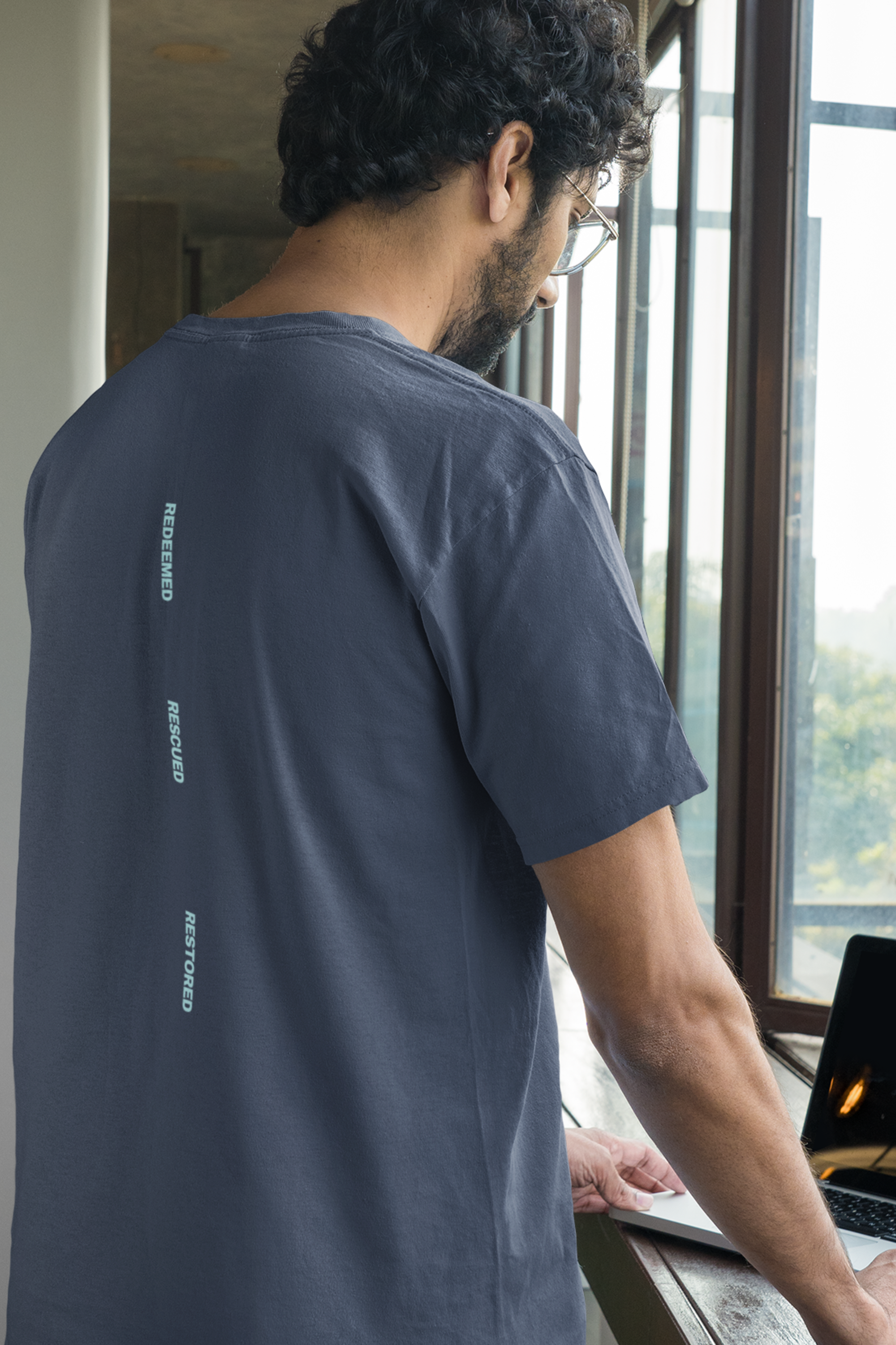 Man wearing a navy blue Christian t-shirt with bible text on the back, standing by a window with a laptop.