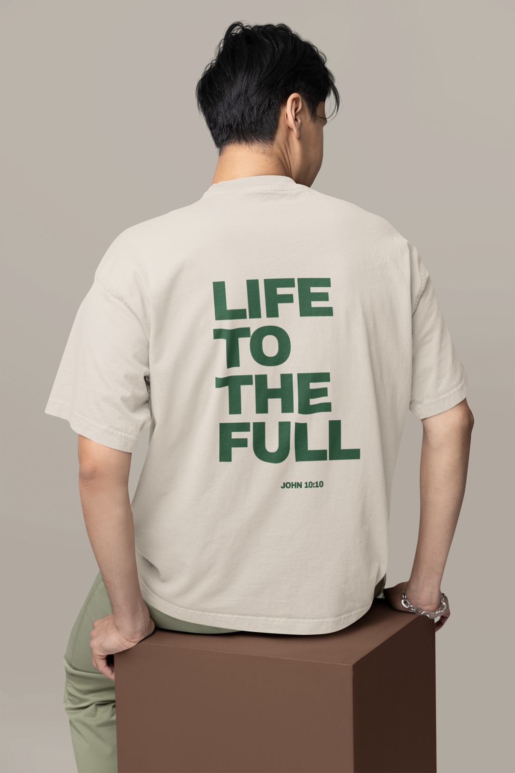 Person wearing a beige t-shirt with 'LIFE TO THE FULL' text on the back, standing on a brown box against a plain background.