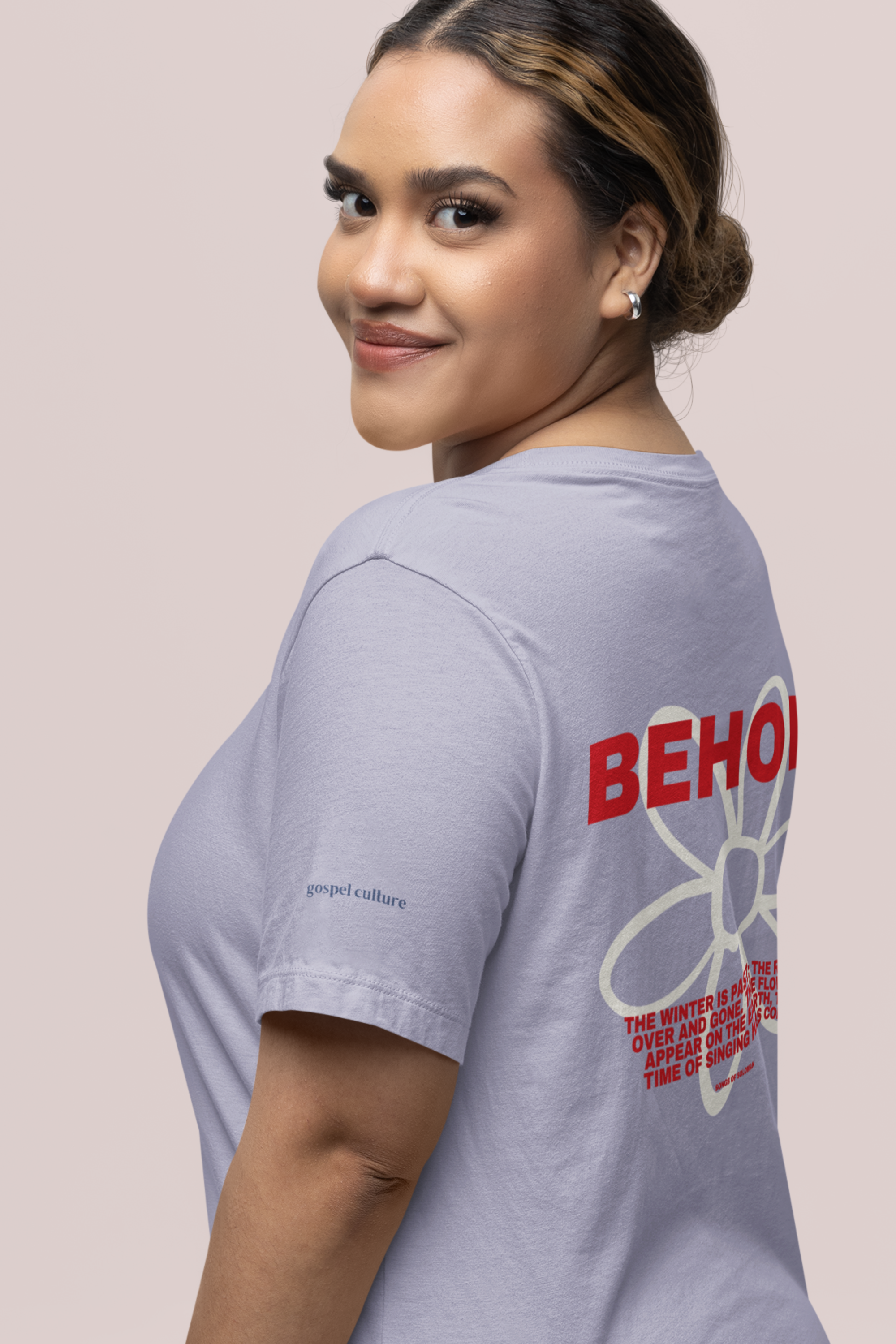 Woman wearing a lilac t-shirt with 'BEHOLD' design on a light pink background