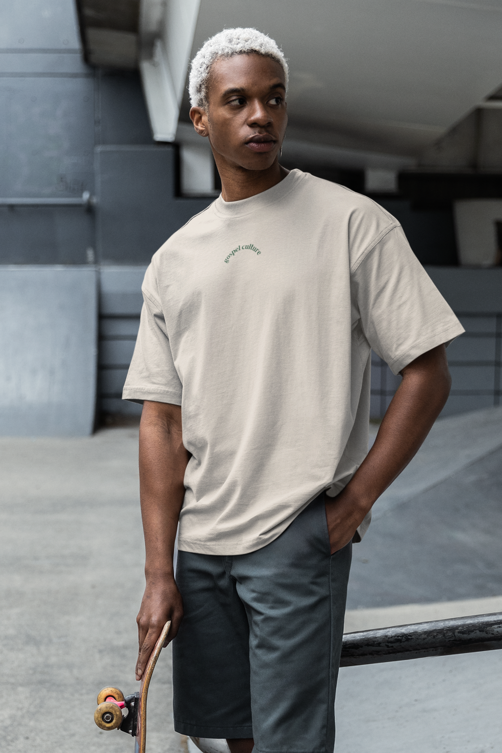Man holding a skateboard wearing a beige  Christian t-shirt and gray shorts in an urban setting.