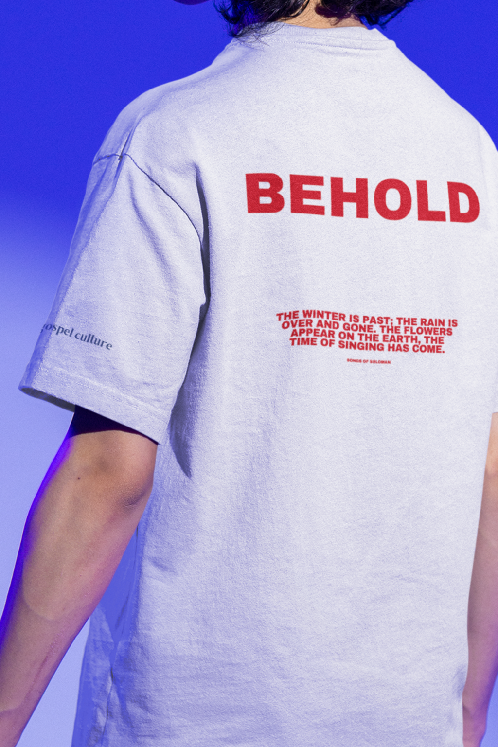 Person wearing a white organic  t-shirt with 'BEHOLD' printed on the back against a blue background