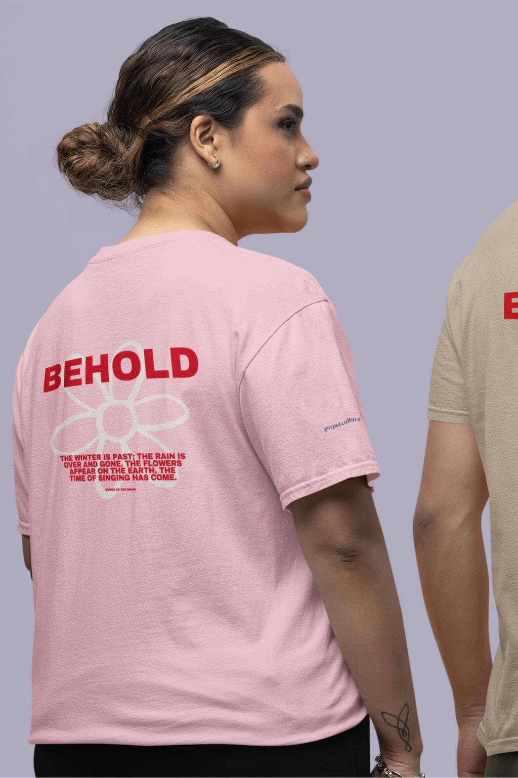 Person wearing a pink t-shirt with 'BEHOLD' text and flower on the back, standing against a plain background.