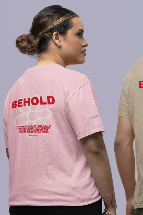 Person wearing a pink t-shirt with 'BEHOLD' text and flower on the back, standing against a plain background.