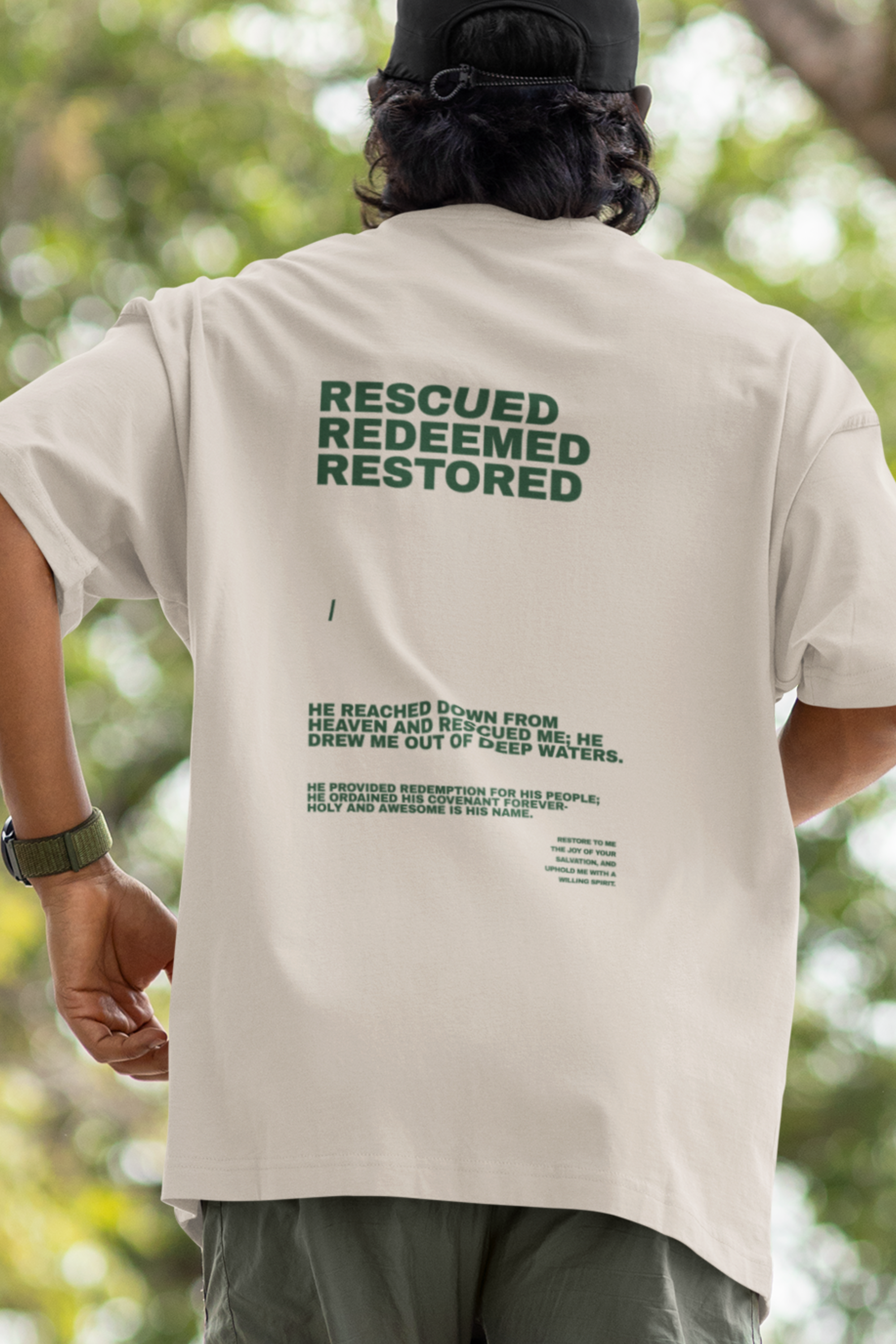 Person wearing a beige t-shirt with 'Rescued Redeemed Restored' text on the back, standing outdoors.