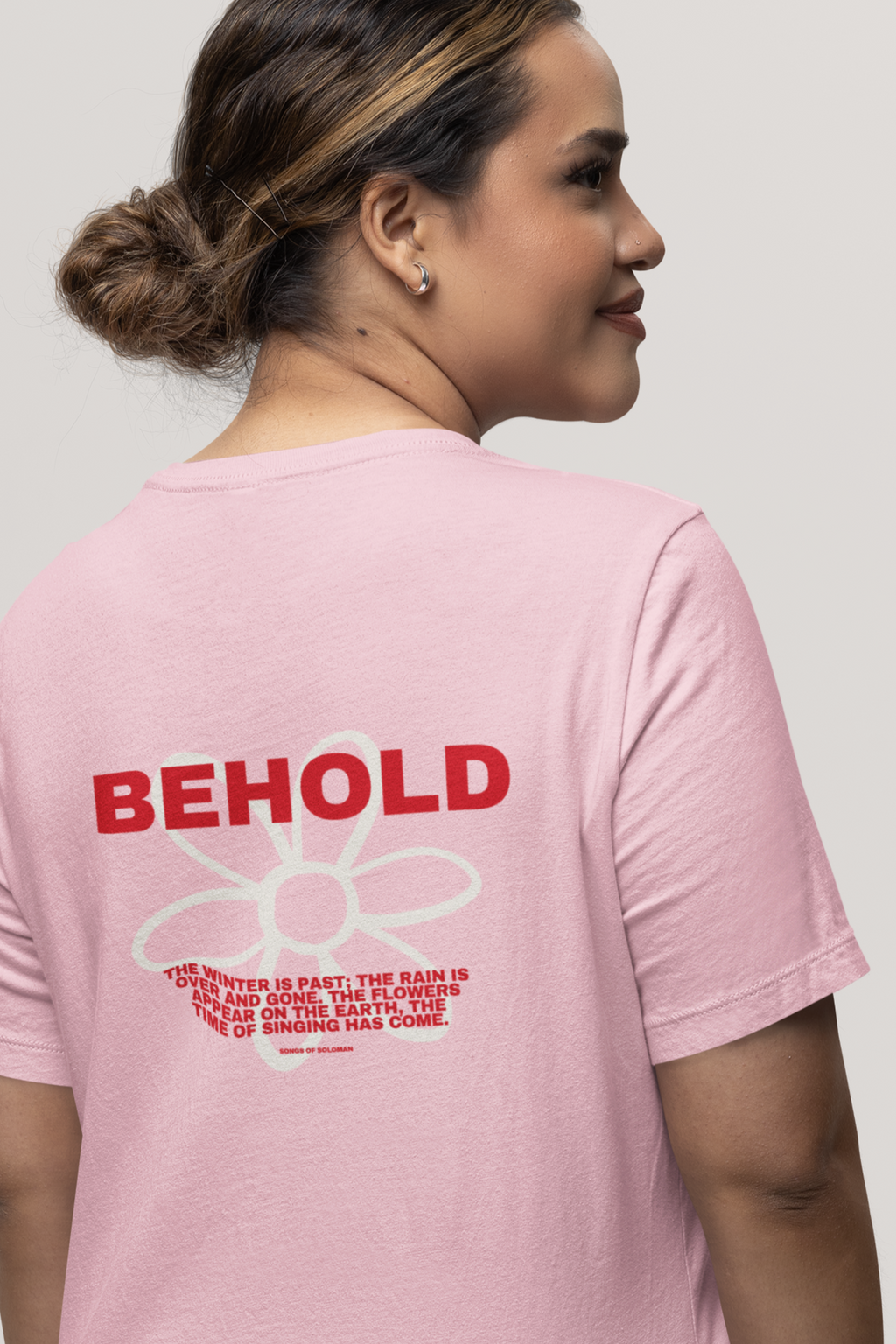 Woman wearing a pink t-shirt with 'BEHOLD' text on the back, against a plain background