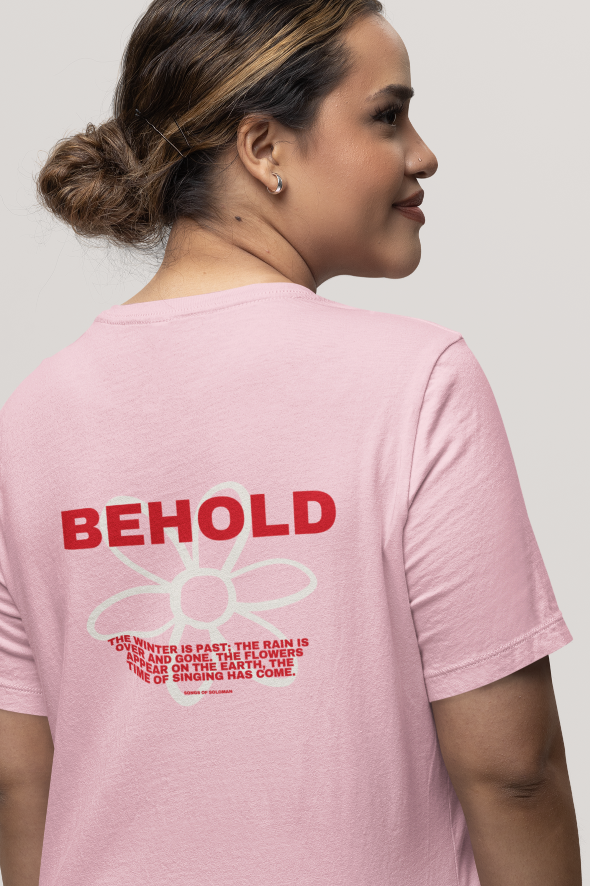 Woman wearing a pink t-shirt with 'BEHOLD' text on the back, against a plain background